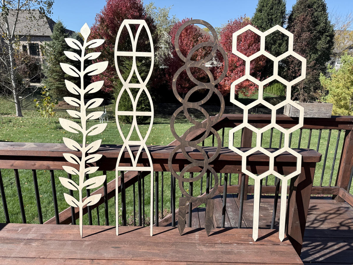 Four laser-cut wood plant trellis designs by Downsized Designs displayed on a deck, leafy vine, helix, circle, and hexagon patterns in unstained Baltic birch