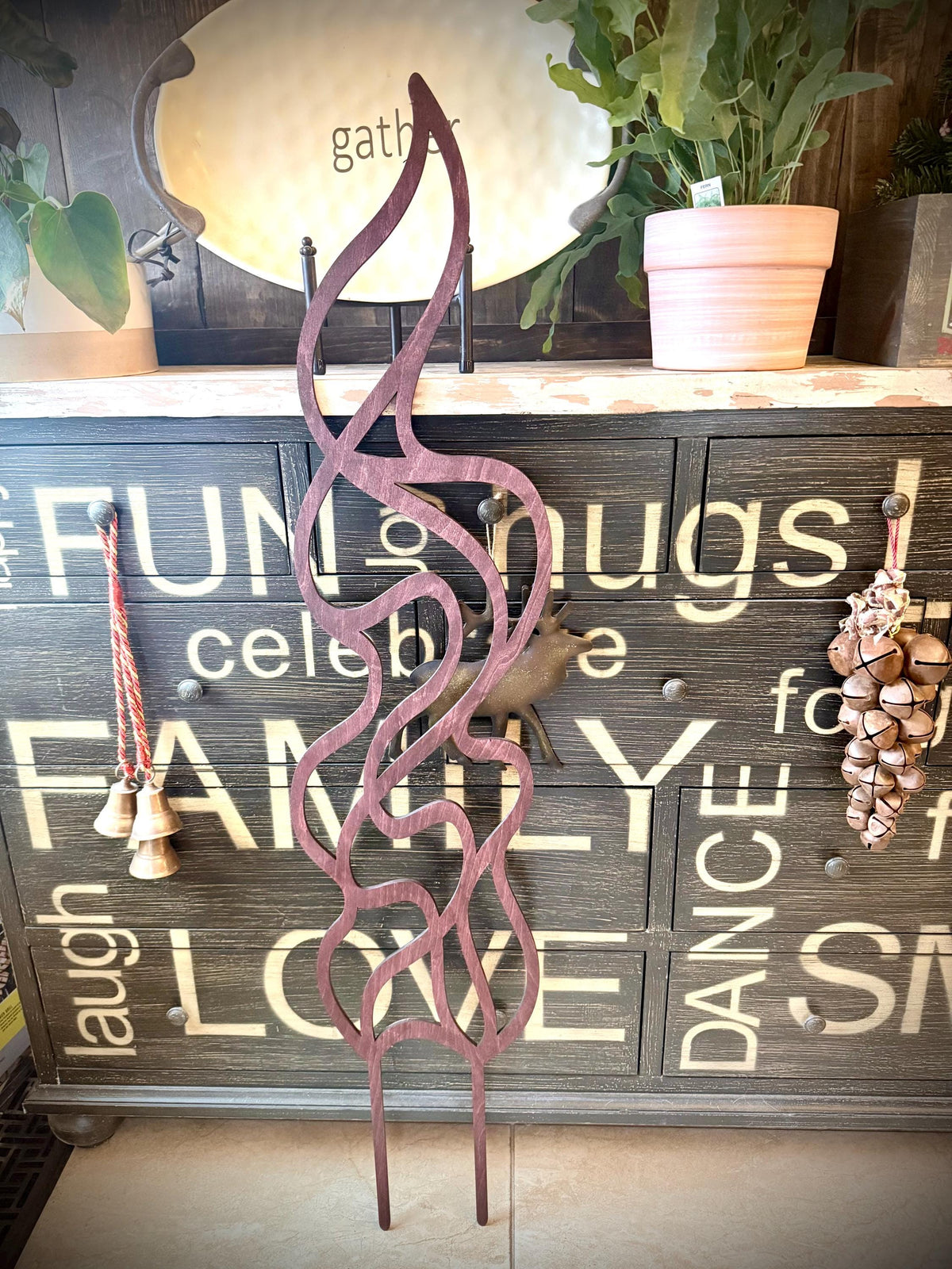 Flame-shaped wooden plant trellis displayed in front of a rustic farmhouse-style chest, with hanging bells and ornaments and a potted plant in the background.
