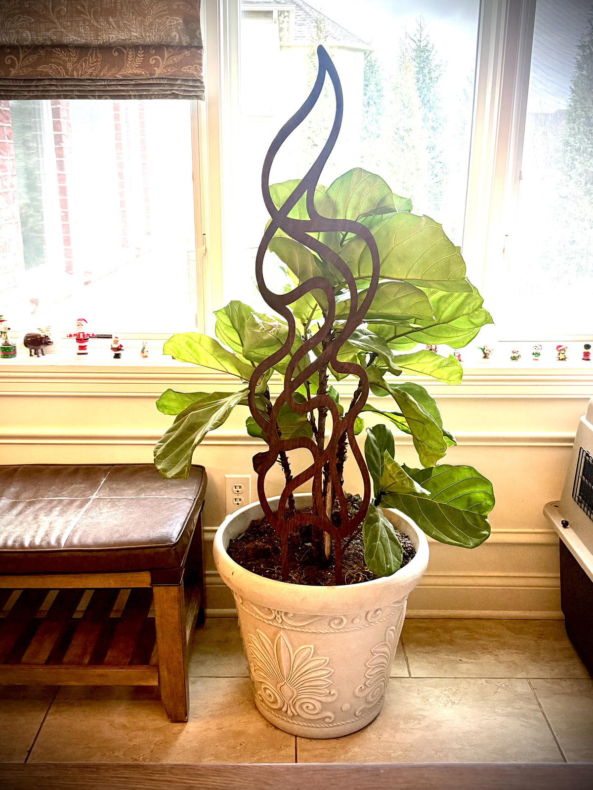 Flame-shaped wooden plant trellis supporting a leafy houseplant in an ornate white pot in front of a bright window, beside a wooden bench.