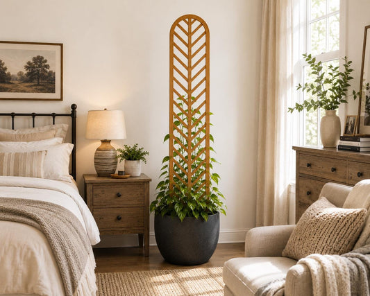 Colossal Herringbone indoor plant trellis in golden oak finish supporting a climbing plant in a black round pot, styled in a warm farmhouse bedroom with linen bedding and wood nightstand