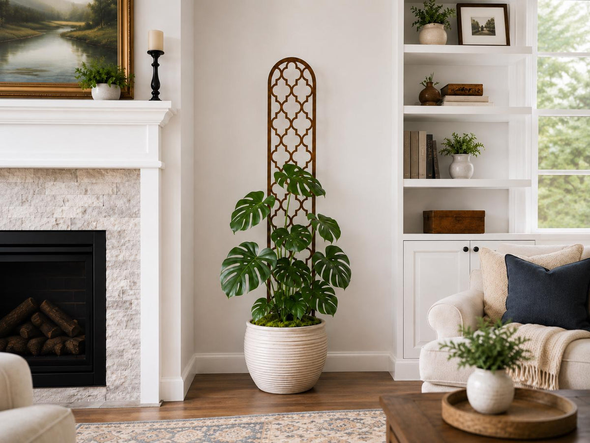 Tall quatrefoil wooden trellis supporting a large monstera plant in a white ceramic pot, displayed next to a fireplace in a classic white living room