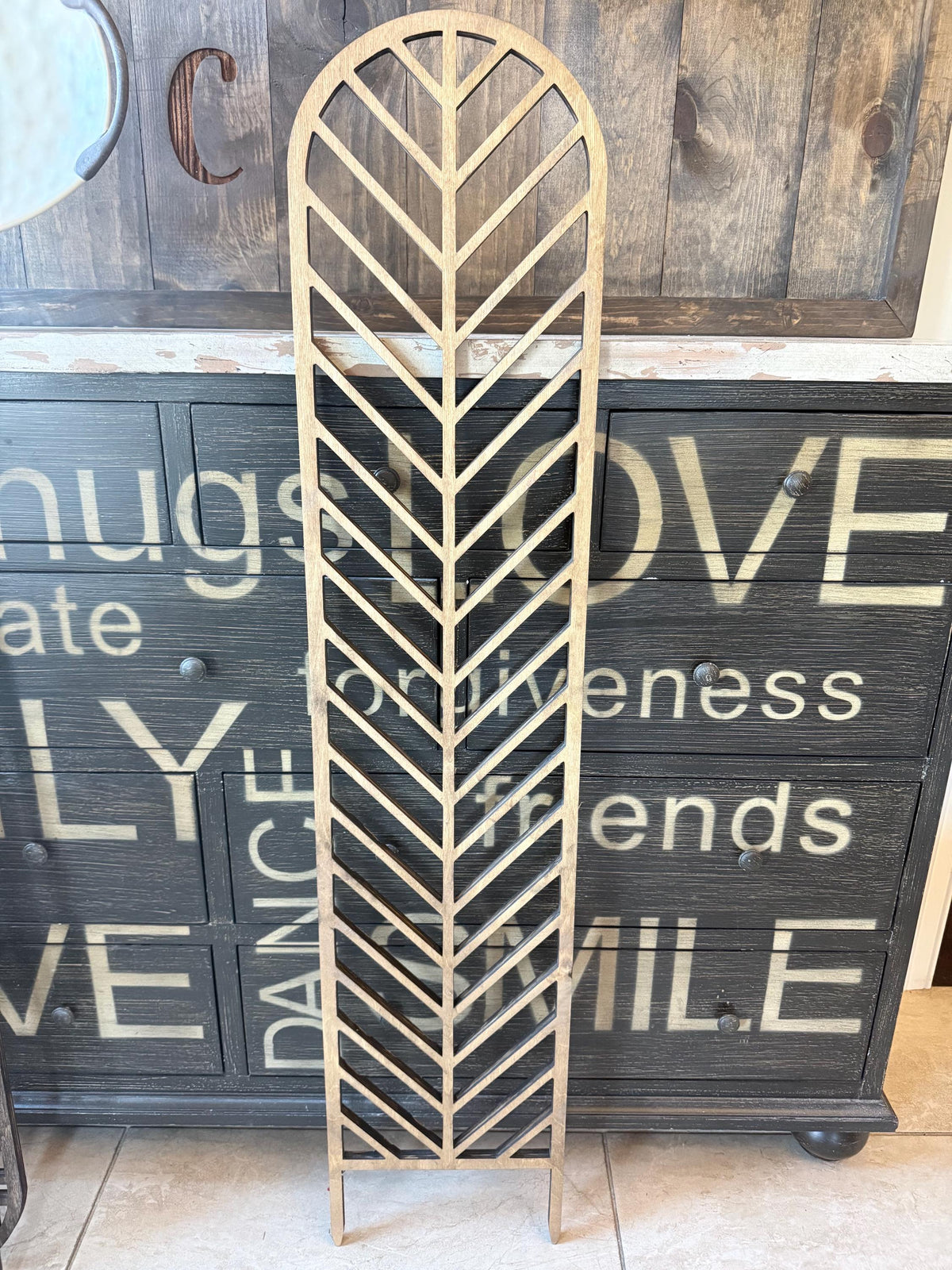 Colossal Herringbone indoor plant trellis in a light wood finish leaning against a rustic wood cabinet